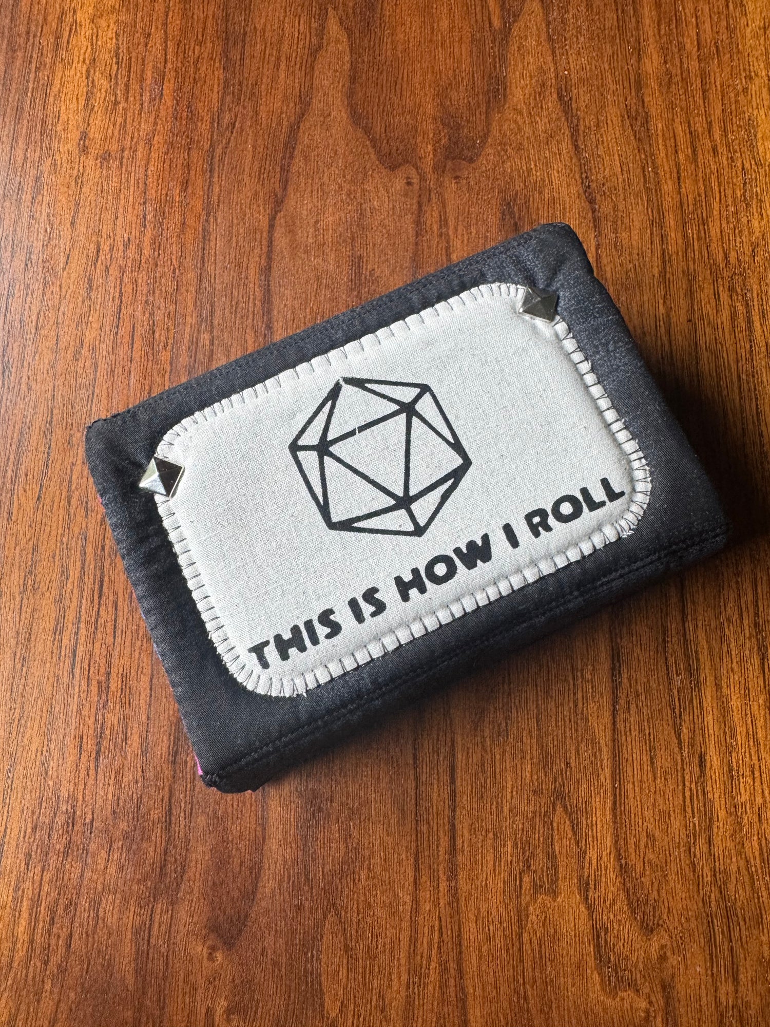 A dark gray fabric bifold wallet with a light gray rectangular patch on the front. The patch features a black line drawing of a 20-sided die and the text 'THIS IS HOW I ROLL' in black capital letters.