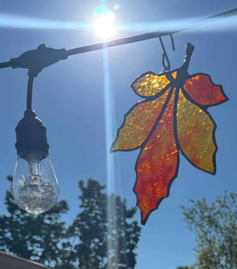 A decorative hanging leaf-shaped stained glass ornament with black metal outlines, shown backlit by a bright sun against a blue sky. A hanging light bulb is also visible on the left, and blurred trees appear in the background.