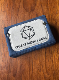 A blue fabric zippered pouch with a white rectangular patch on the front featuring a black d20 die icon and the text 'THIS IS HOW I ROLL', photographed on a wooden surface.