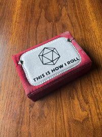 A red and pink fabric dice rolling pouch with a white patch that reads 'THIS IS HOW I ROLL' and features a black d20 die icon, resting on a wooden table.