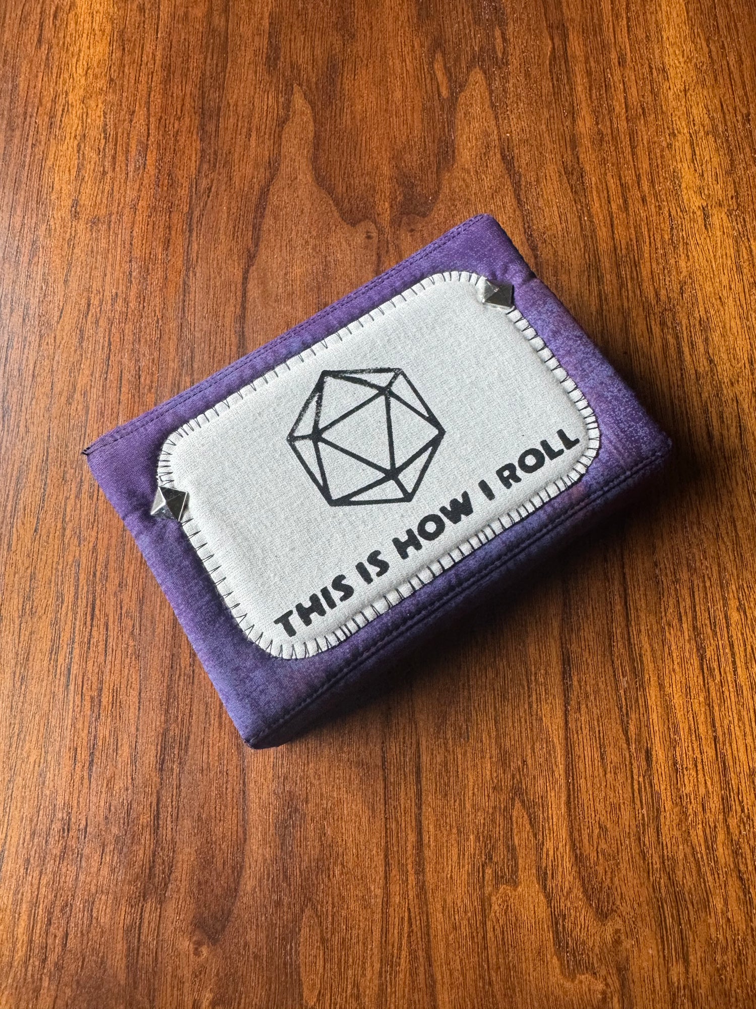 A purple fabric card holder or wallet with a white stitched patch on the front that reads 'THIS IS HOW I ROLL' with a geometric d20 die icon, placed on a wooden surface.