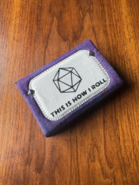 A purple fabric card holder or wallet with a white stitched patch on the front that reads 'THIS IS HOW I ROLL' with a geometric d20 die icon, placed on a wooden surface.