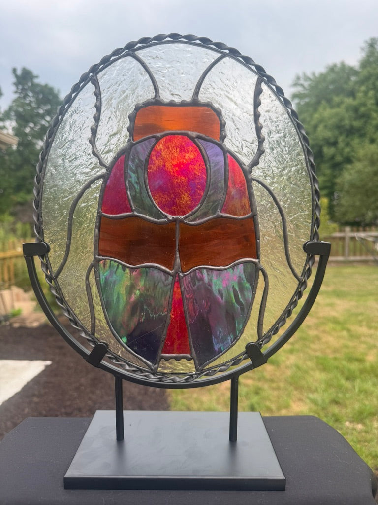 A stained glass scarab design mounted on a black metal stand, set against a blurred outdoor background of green grass and trees.