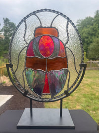 A stained glass scarab design mounted on a black metal stand, set against a blurred outdoor background of green grass and trees.